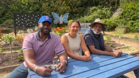 Three people are sitting on a blue garden fence looking into the camera and smiling. 