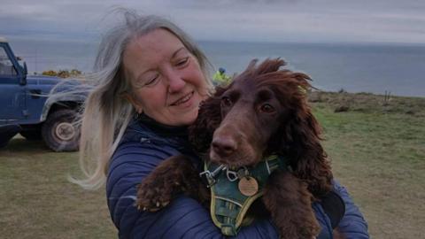 A cocker spaniel in the arms of a women with long grey hair on a grassy cliff top