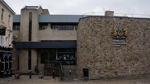 The front entrance of Plymouth Magistrates' Court on a cloudy day. A large sign for the court is on a brick wall. A pub is to the left of the court building.