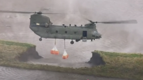 Large helicopter drops off heavy bags to stop flooding that has created a huge hole in a grassy bank.