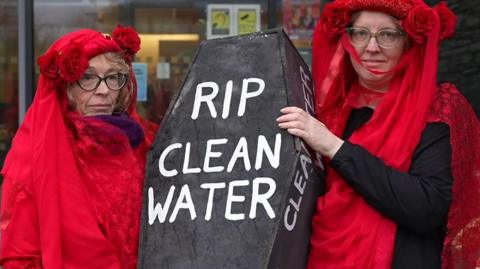 Two protesters are wearing red and are holding a coffin that says RIP clean water.