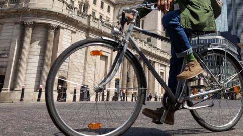 A man in blue jeans, a green coat and brown shoes on a bicycle in a city