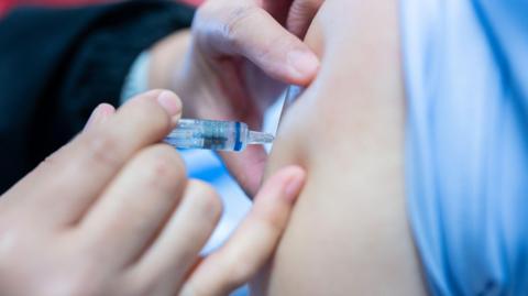 A nurse is giving a person a jab. The sleeve is rolled up above the person's arm and the nurse is pressing a needle into the arm.