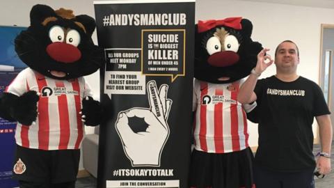 Michael Chapplow is wearing a black t-shirt reading ANDYSMANCLUB. He is standing next to banners about encouraging men to talk about their mental health. Two Sunderland AFC black cat mascots in the club's red and white t-shirts are standing on each side of the banner.