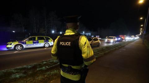 A police officer wearing a flak jacket with police written across the back. They're also wearing a bright yellow jacket underneath with police branding on it. A police car can be seen in the background.