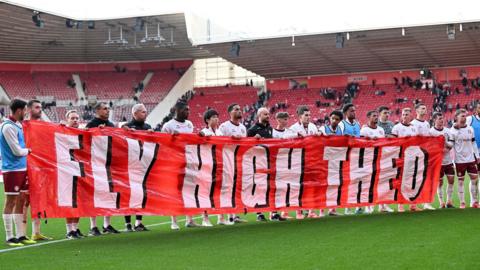 Bristol City players lined up on the football pitch holding a banner which reads: "Fly high Theo"