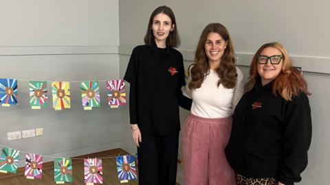 Three women stand side-by-side next to a display of children's artwork. Two of the women are in black The Lewis Foundation branded shirts while Martine Soldano is in a white top and pink trousers.