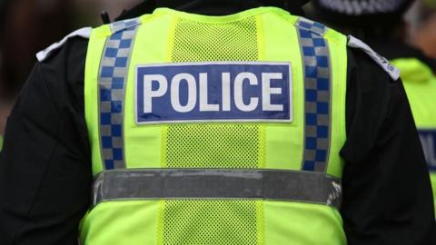 A police officer in a high vis vest with the word 'police' on the back of it stands facing away from the camera. 