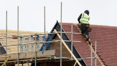 Scaffolding on the outside of two new build houses, roofs only in vision. A man is on top of the roof on the right.