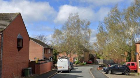 A street view image of a road with houses on each side and cars parked on the road. The homes are made of red brick and there is a white van on the road nearest the camera. A couple of trees are next to the homes.