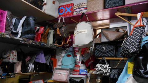 Shelves packed with a large collection of fake designer handbags in various colours and patterns. They are in a small storage room.