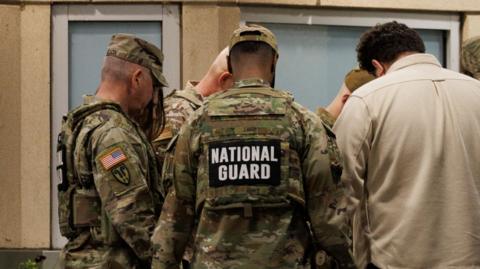 A photograph of a number of National Guard members standing together. At least 5 men can be seen in the image - one of whom appears to be in civillian clothing wearing a cream-coloured shirt. The rest of the men are in uniform and the words 'National Guard' can be seen on the back of their uniform.