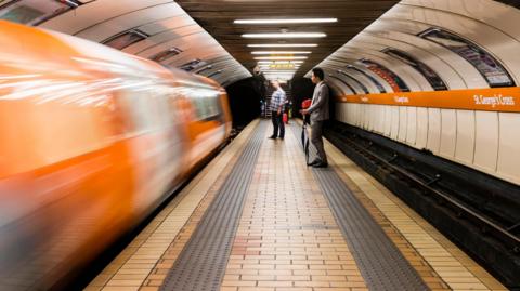 A subway carriage pulling into St George's Cross station blurred. People stand on the platform.