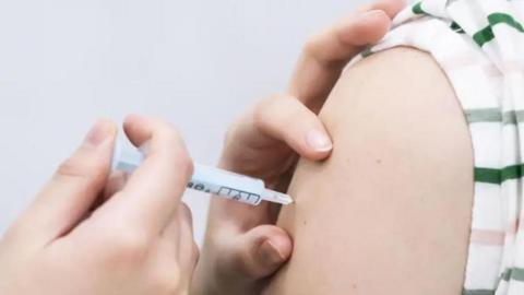 A close-up of a hand injecting a white needle into the top of someone else's arm. The person is wearing a striped green and pink t-shit. 