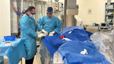 Two surgeons dressed in blue gowns standing over a table in an operating theatre which is covered in a blue sheet