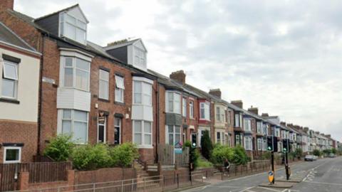A row of terraced housing is on the left hand side of the road. There is a pedestrian crossing with traffic lights in the centre of the road.
