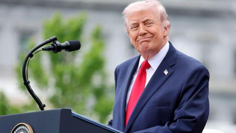 President Trump smiling in front of a microphone. He is wearing a navy suit, red tie and white shirt.