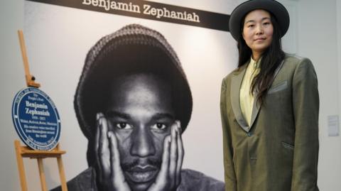 Qian Zephaniah wearing a brimmed hat and green-grey jacket in front of a portrait of Benjamin Zephaniah and blue memorial plaque at a recent event.