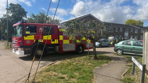 A fire engine parked on a road near an old people's home. It is next to a small tree. It is a sunny day.