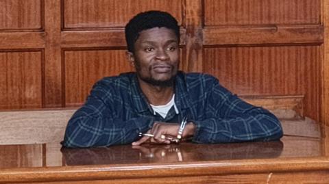 Webster Ochora Elijah in a checked shirt, behind the dock in court