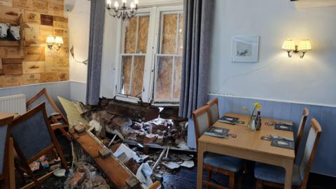 A pile of tiles and debris on the floor of the pub. A number of chairs have been pushed down on the floor by the force of the collision. The windows are also pushed in and have been boarded up. There are cracks in the wall. A table with four chairs and a vase with daffodils to the right of the windows remains intact.