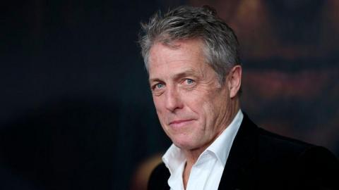 Hugh Grant, who wears an open-necked shirt and suit jacket and has greying hair, looks in the direction of the camera, against a black background, at a premiere.