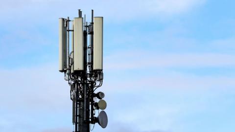 The top of a vertical mobile mast. It's a black pole covered in wires and communication technology. There's a blue sky in the background.