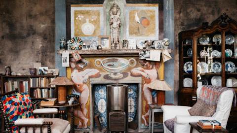 A small art studio centred around a fire place with two arm chairs either side facing on another. There is a book case to the left of the fire place and a large plate cabinet to the right. There a small pieces of art on the mantle piece.