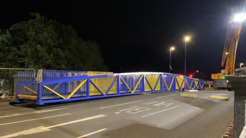 A long blue and yellow painted footbridge resting on a road bridge at night, ready to be lifted into place. There is a yellow crane in the background.