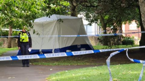 A forensic tent in a woodland area with a police cordon around it and a police officer in a black uniform and a hi-vis green jacket standing by the tent