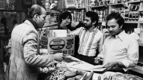 Black and white photo of shopkeeper Komor Uddin serving a customer at his grocer's shop, the Taj Stores in Brick Lane