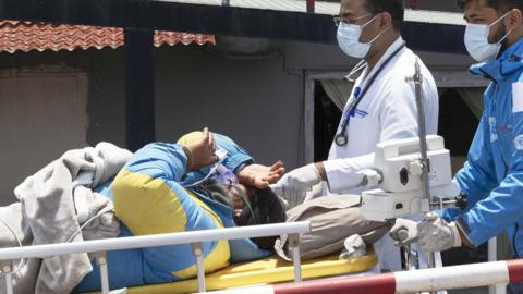 Dawa Sherpa on a stretcher acompanied by two doctors. He is wearing a blue and yellow mountian jacket