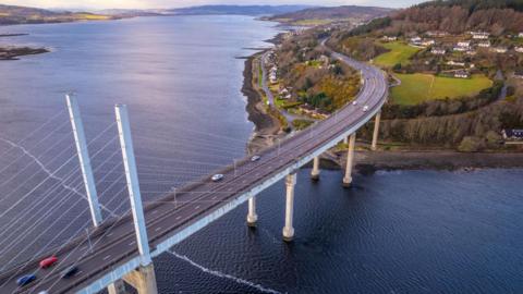 An aerial image of the Kessock Bridge with the Beauly Firth and Inner Moray Firth on either side of the crossing on the A9 trunk road. On the area of headland in the shot are white-walled homes of North Kessock.