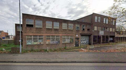 A two-storey building, seen from the outside and with graffiti on the downstairs windows, is most prominent in the photo. The pavement and part of a road is in the foreground.