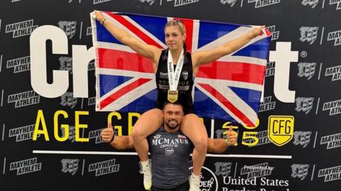 Keira, sat on her dad Joel's shoulders, is holding a union jack flag and has a medal around her neck. They are in front of a step and repeat board, with the word Crossfit and various sponsorship logos on it.