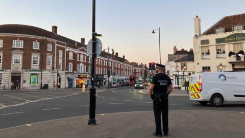 A police officer stands with his back to the camera on a street corner in Epsom. The road visible in front of him has three storey buildings along it with shops at ground floor level, and it is early evening.