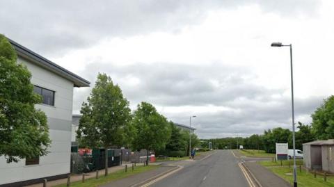 A road with large industrial units either side. There are double yellow lines and junctions leading to units on the left hand side and right hand side of the road.