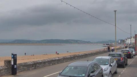 Image shows Exmouth seafront on a grey day. A number of cars are parked in the bays on the left-hand side of the image. People are walking along the esplanade behind the beach. There is a stone wall separating the walkway and the sandy beach. A number of people are standing on the sand. 