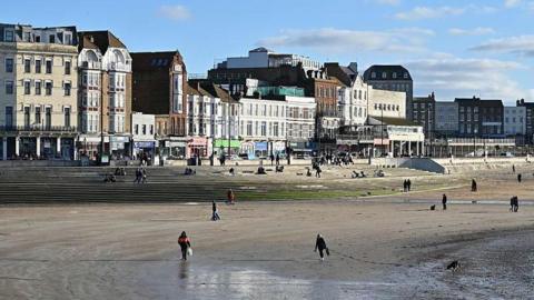 Properties by a seafront. There are people on the beach, some walking their dogs.