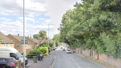 Hylton Road in Worcester with a wall on one side and houses on the other, with cars and vans parked outside. There are trees, ivy and shrubs overhanging the wall and there are cars driving in the road. A woman is walking on a pavement near a wheeled bin.