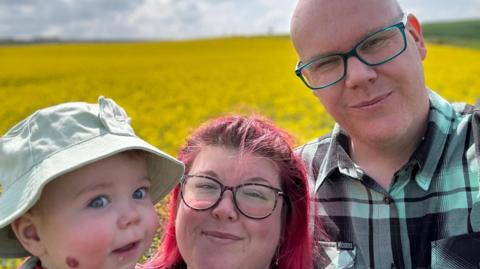 A selfie of Tim Brentnall, his wife and child. Tim is wearing a green and black checked shirt, and green glasses. His wife has red hair and is wearing dark glasses. The toddler is wearing a light coloured hat. They all look at the camera. 