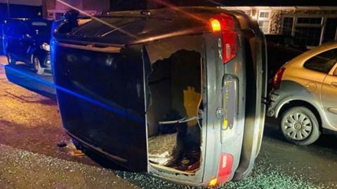 A car turned over on its side in the middle of a residential street. It is a dark colour and the hazard lights are on. The back windscreen is fully smashed, with some items visible in the boot. It is dark and there are houses and parked cars in the background. Blue lights from a police car can be seen on the left. 