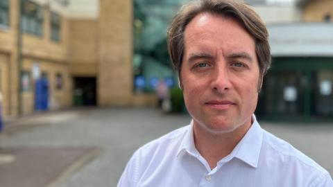 Thom Lafferty stands outside the newer front entrance of the Princess Alexandra Hospital in Harlow and looks down the barrel of the camera. It has beige brick and a glass staircase. He is wearing a white collared shirt and has blue eyes and brown hair