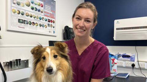 Amy Wheeler, a young woman wearing purple scrubs, smiles as she sits in a vet's office. Her dog Rona is sitting on her lap facing the camera.