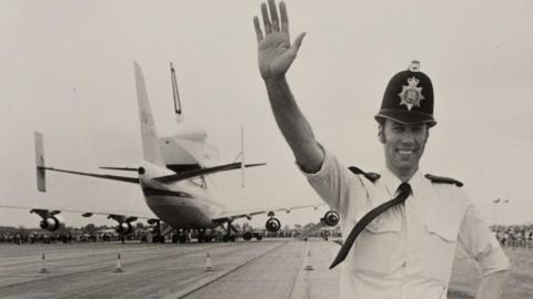 A black and white photo of officer Brian Bishop. He is pictured in police uniform at Stansted Airport. He has one arm up in the air and the other resting on his hip. He is smiling to the camera and an aeroplane is parked behind him.