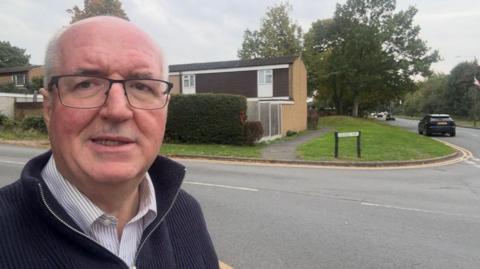 A man wearing a blue jumper and a white pin-striped shirt, stood in a residential street in Droitwich.