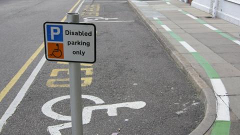 A disabled parking bay with markings painted on the road and a sign on a short metal pole. There is a wheelchair drawn on the road and on the sign.