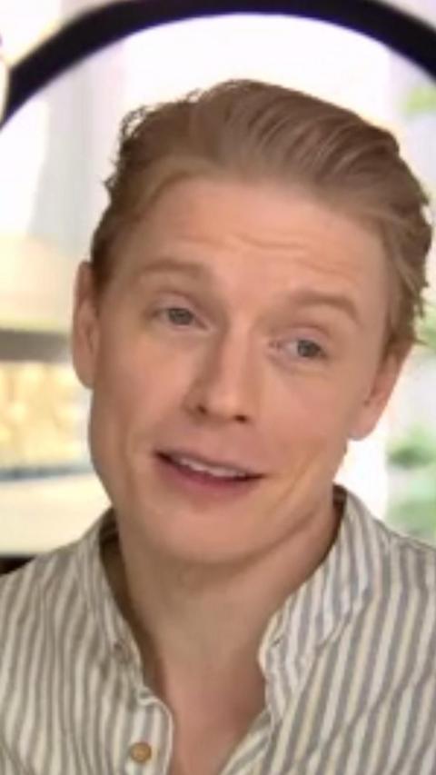 Freddie Fox has blonde hair swept back, he is wearing a grey and white striped collared shirt.