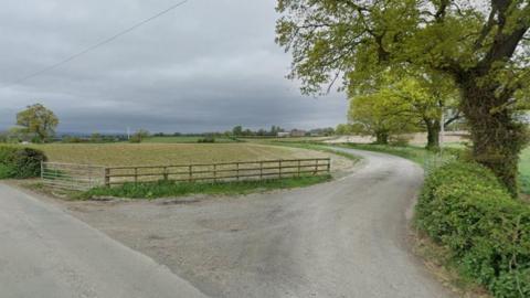 A winding road leads to a fam building in the distance. It is surrounded by green fields and trees. The sky is grey.