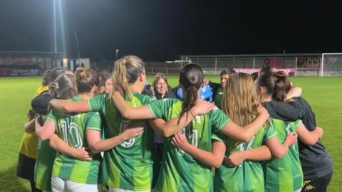 The Lionesses form a circle on a football pitch at night with floodlights on and with their arms around each other 